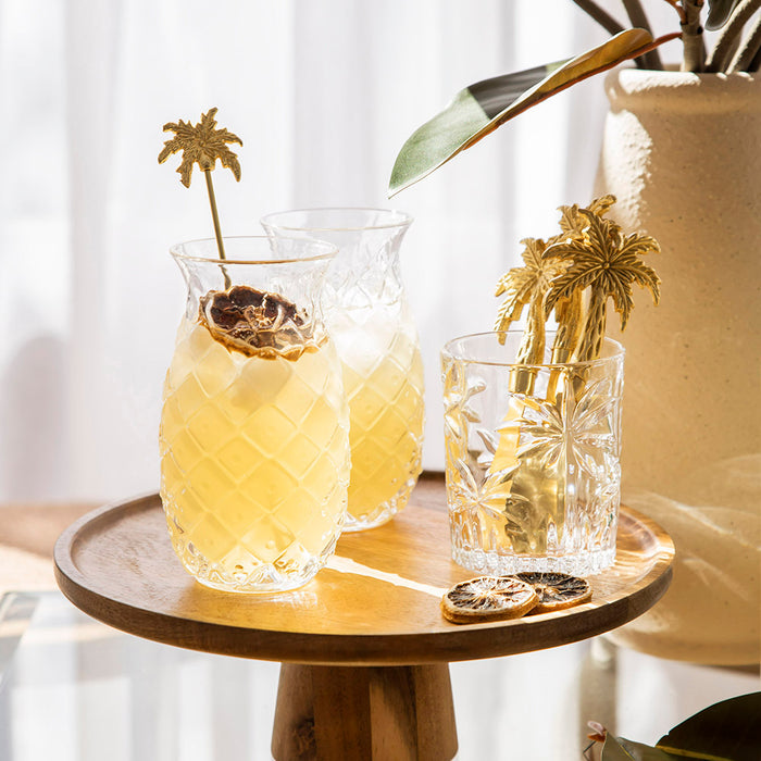 Serving plate featuring two cocktails in pineapple shaped glasses and a tumbler glass holding gold palm tree shaped utensils.