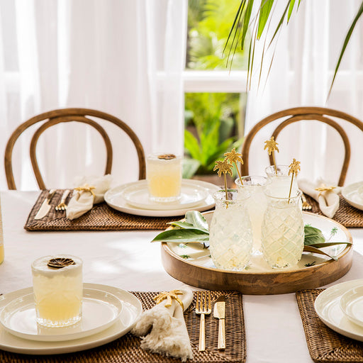 Fully set dining table featuring four cocktails in pineapple shaped glasses with gold palm tree drink stirrers. 